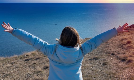 The girl stands facing the sea with her arms outstretched to the sun, March 2023